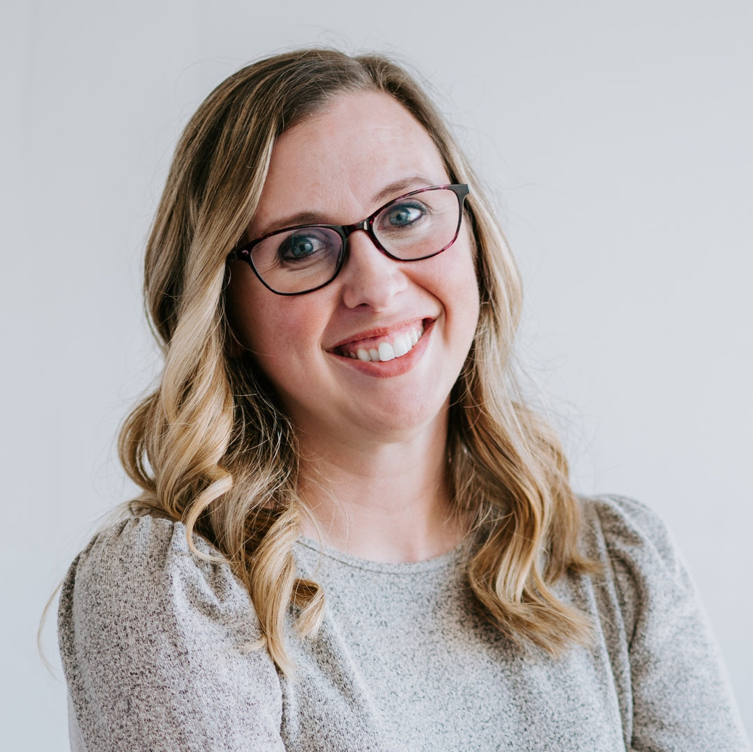 Jessica Deen Norris - A smiling woman with blonde hair and glasses wearing a light gray textured top, photographed against a plain light gray background.